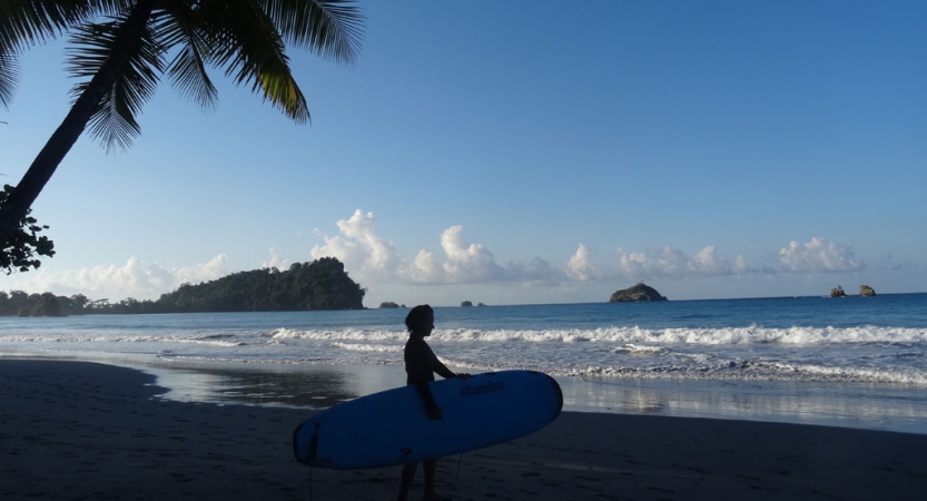 A person stands on a beach holding a surfboard. There are small islands in the distance. 